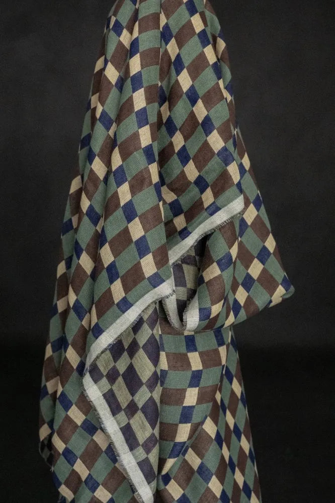 A draped fabric item, likely a shawl or scarf, featuring a geometric diamond pattern in olive green, brown, navy blue, and cream, with a contrasting light gray border, set against a dark background.