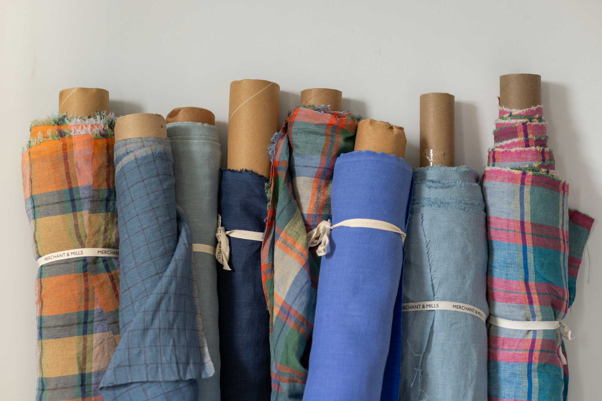 A group of rolled fabric bolts secured with white bands, standing upright against a plain light wall. The fabrics show various patterns and colors including orange, blue, green, pink, and plaid designs.
