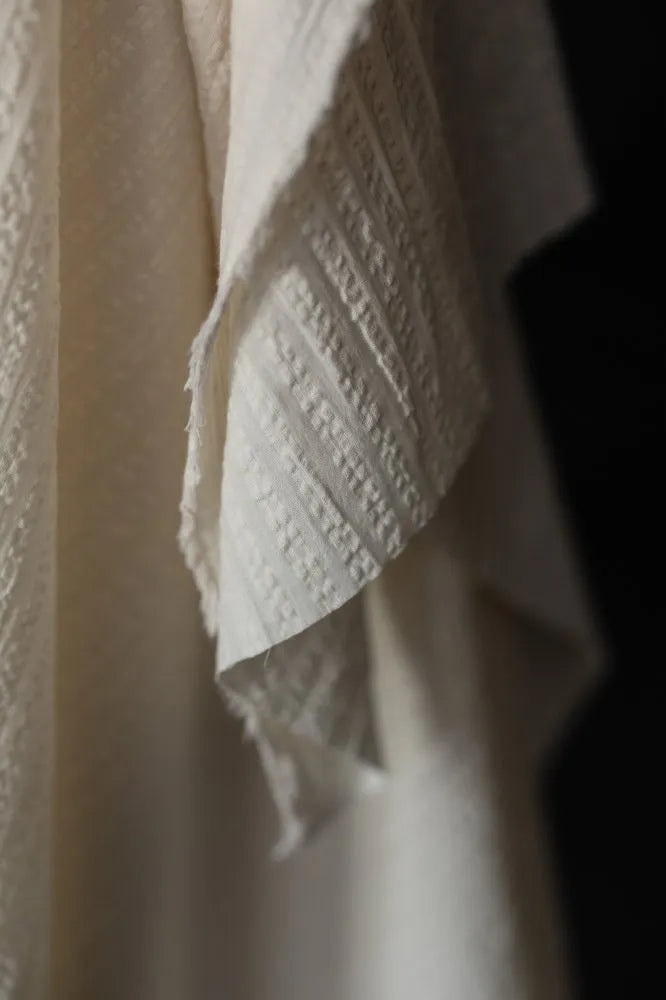 A close-up of draped off-white textured fabric with a woven basketweave pattern, set against a dark background.