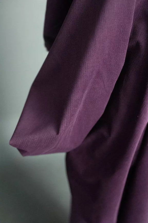 A close-up of a draped purple garment, showing the fabric texture and folds against a soft gray background.
