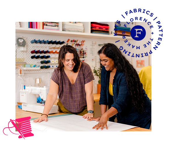Two women collaborating in a sewing room, looking at a large sheet of white fabric on a table. A white sewing machine is visible in the background, along with shelves of thread and fabric spools.