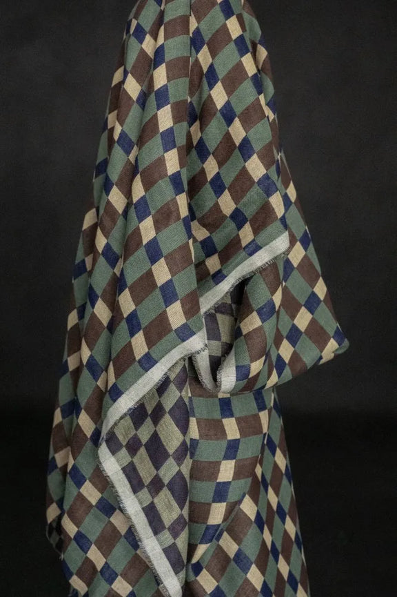 A draped fabric item, likely a shawl or scarf, featuring a geometric diamond pattern in olive green, brown, navy blue, and cream, with a contrasting light gray border, set against a dark background.