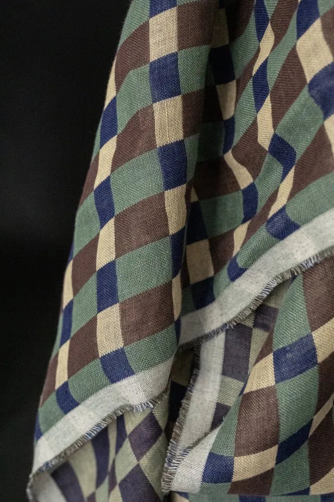 A close-up of a woven scarf or fabric with a geometric diamond pattern in olive green, navy blue, brown, and beige, featuring a frayed edge.